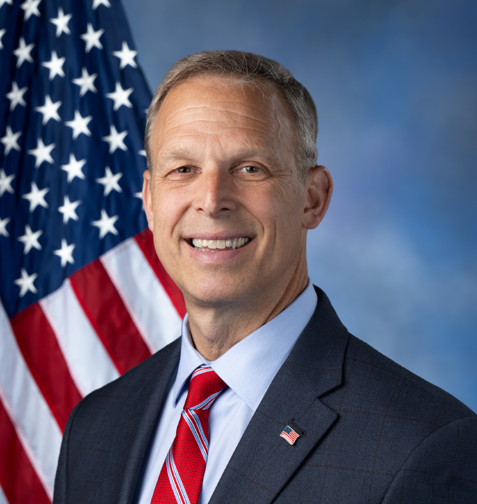 A man in a suit and tie standing in front of an American flag, smiling.