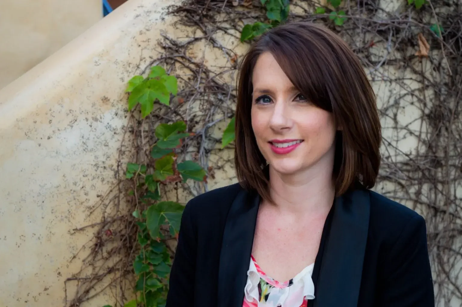 A woman with shoulder-length brown hair, blue eyes, and pink lipstick, wearing a black blazer and a floral top, standing outdoors in front of a wall covered with green ivy and vines.