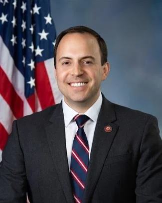 A man in a dark suit, white shirt, and striped tie standing in front of American flags, smiling.