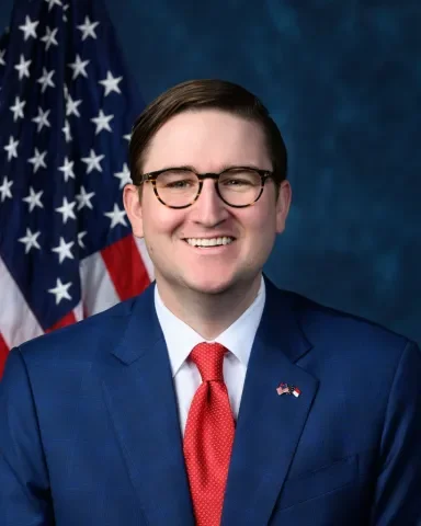 A man in a blue suit, red tie, and glasses, smiling in front of an American flag backdrop.