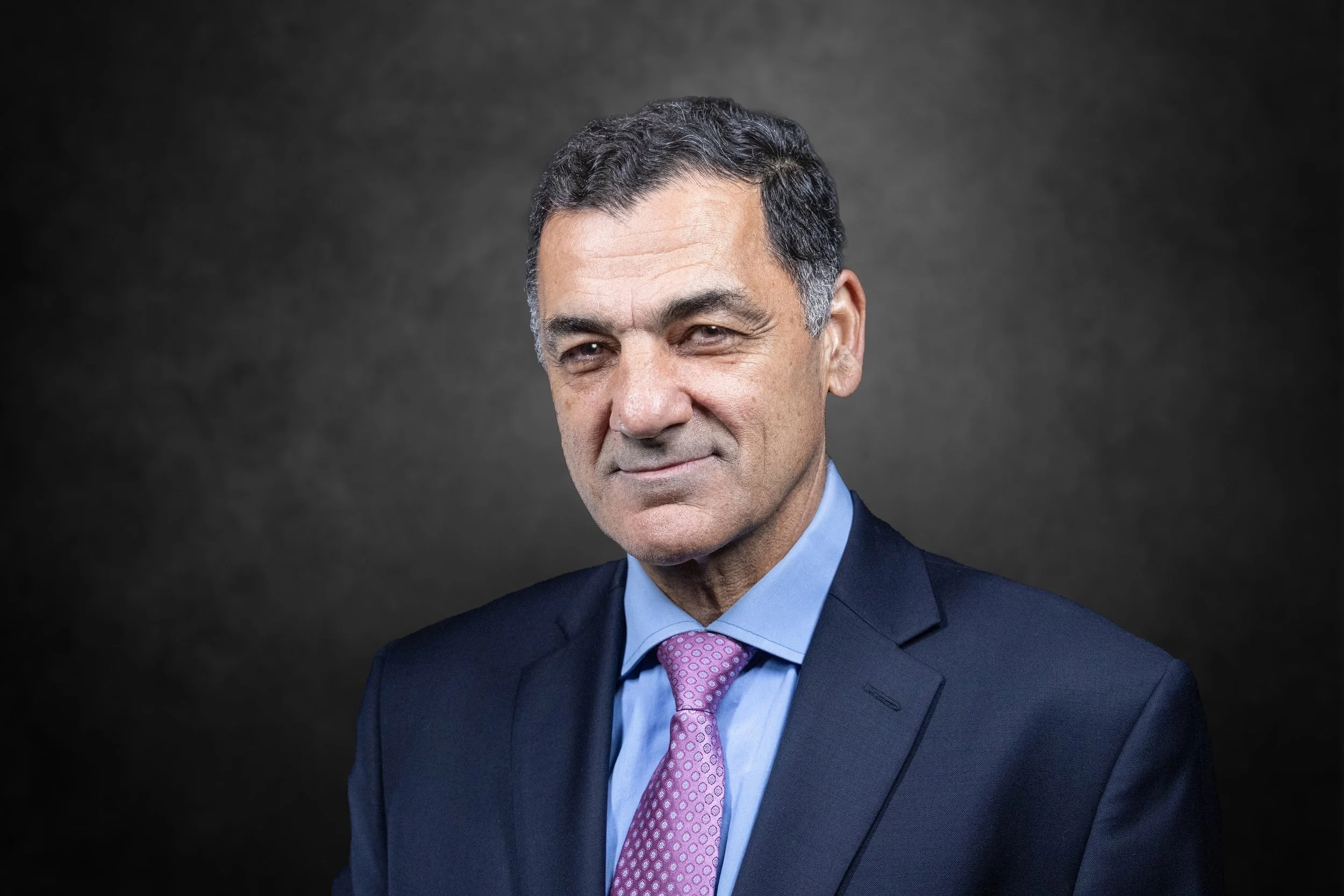 A middle-aged man in a business suit with a blue shirt and pink tie, looking at the camera with a slight smile against a dark gray background.