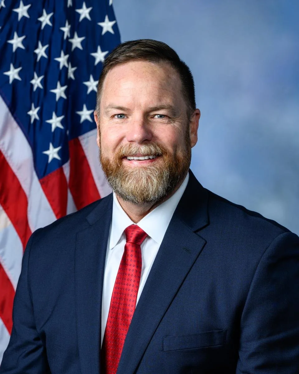 Portrait of a man with a beard and short hair, wearing a navy suit, white shirt, and red tie, standing in front of an American flag with a blue sky background.