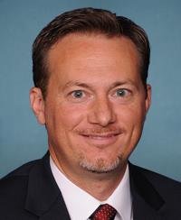 A professional headshot of a middle-aged man with short brown hair, a goatee, wearing a dark suit, white shirt, and red patterned tie, against a plain background.