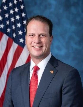 A man in a suit and red tie standing in front of an American flag.