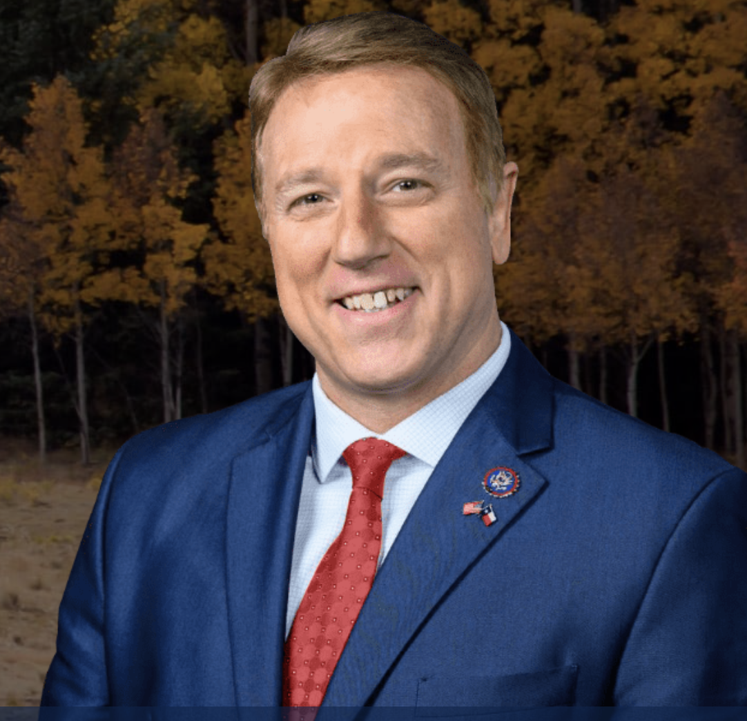 A smiling man in a blue suit with a red tie and patriotic pins, standing outdoors in front of a background of fall-colored trees.