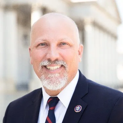 A smiling man in a suit outdoors, with a white shirt, a striped tie, and a lapel pin, blurred buildings in the background.