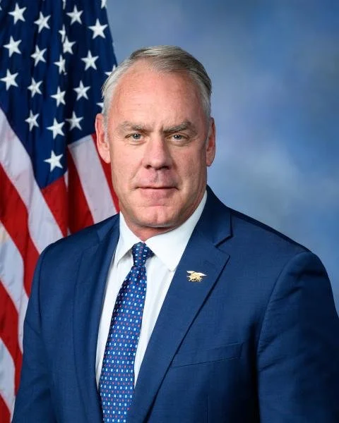 A middle-aged man in a blue suit, white shirt, and patterned blue tie, standing in front of the American flag against a blue background.
