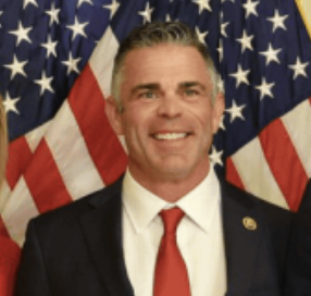 A man wearing a black suit, white shirt, and red tie, standing in front of American flags, smiling.