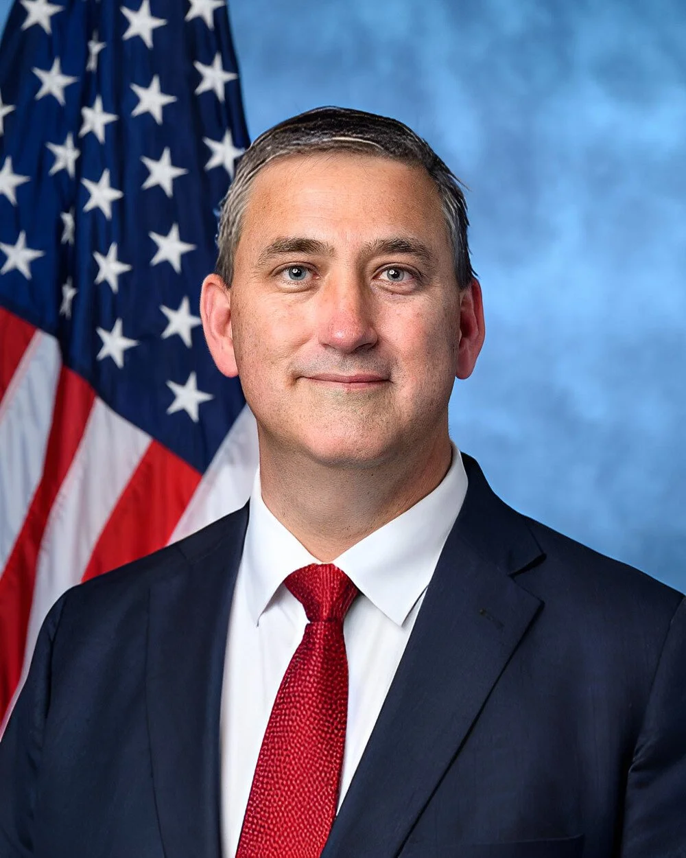 A man in a suit and red tie standing in front of an American flag with a blue background.