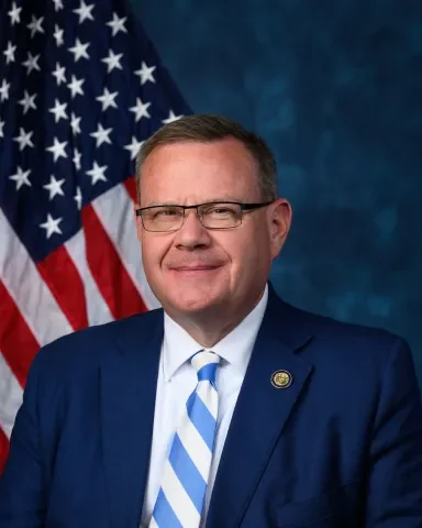 Portrait of a man in a dark blue suit and tie, wearing glasses, standing in front of an American flag.