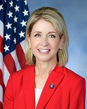 Portrait of a woman with blonde hair, smiling, wearing a red blazer and a white top, standing in front of an American flag.