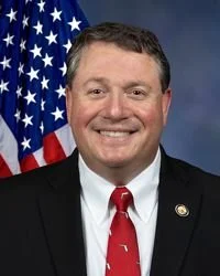 A man in a suit and red tie, smiling, standing in front of an American flag.