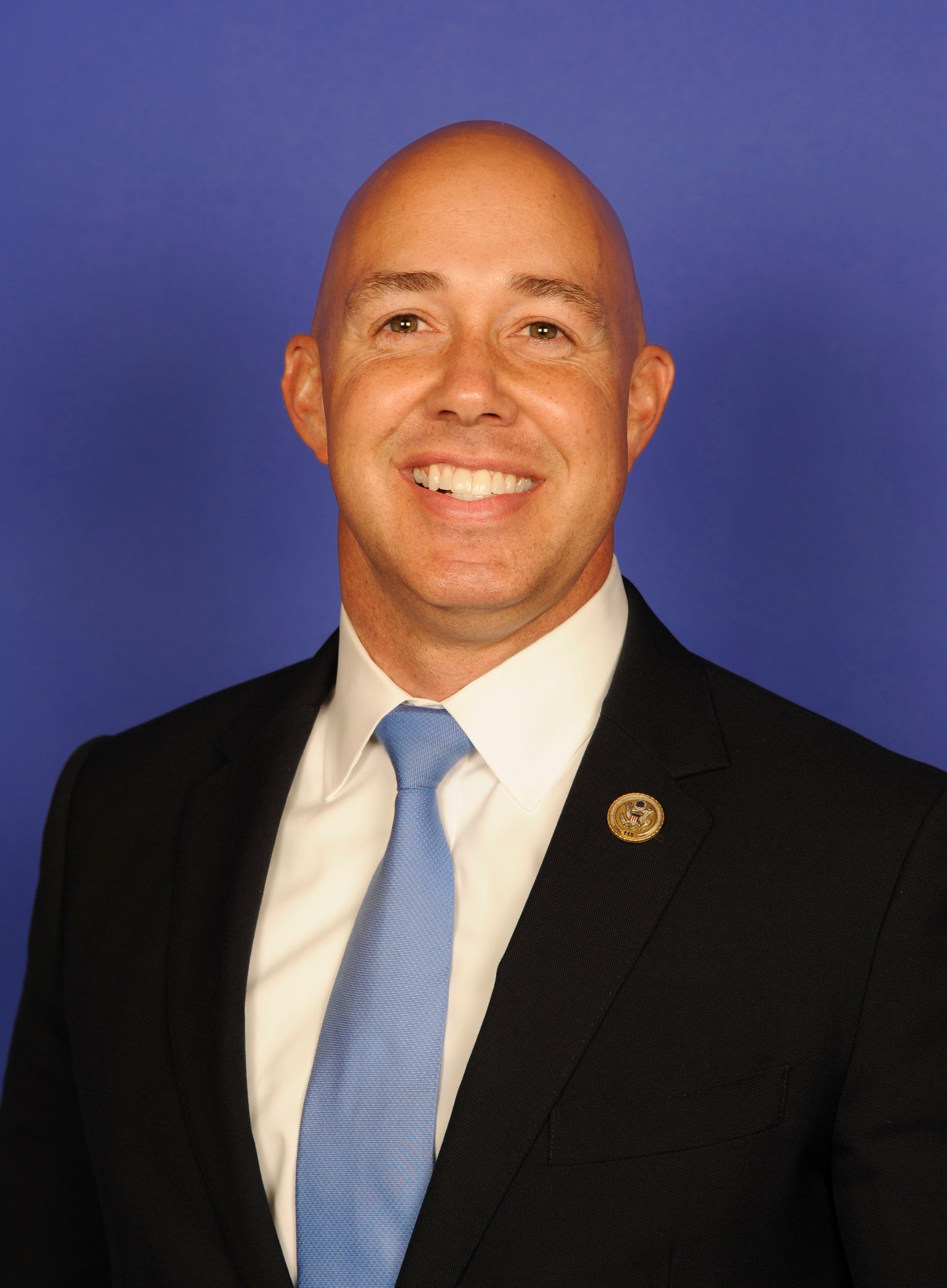 A smiling bald man wearing a black suit, white shirt, light blue tie, and a lapel pin, against a blue backdrop.
