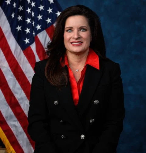 A woman with dark brown hair and a red blouse, wearing a black blazer, standing in front of an American flag and a blue background.