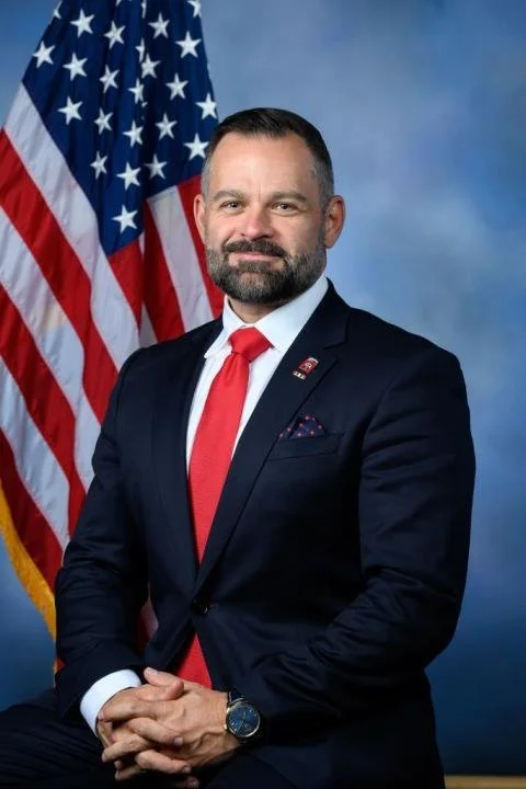 A professional man in a dark suit, white shirt, and red tie sitting in front of an American flag with a blue background.