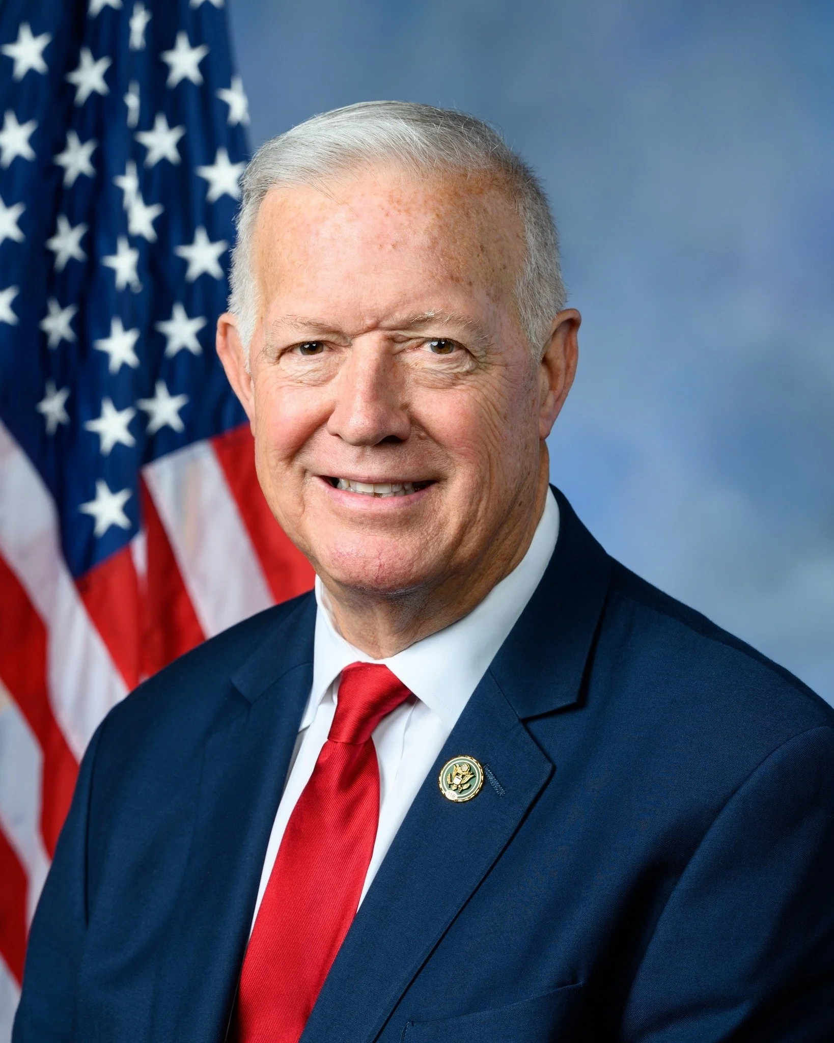 Close-up of a smiling man in a dark suit, white shirt, red tie, with an American flag in the background.