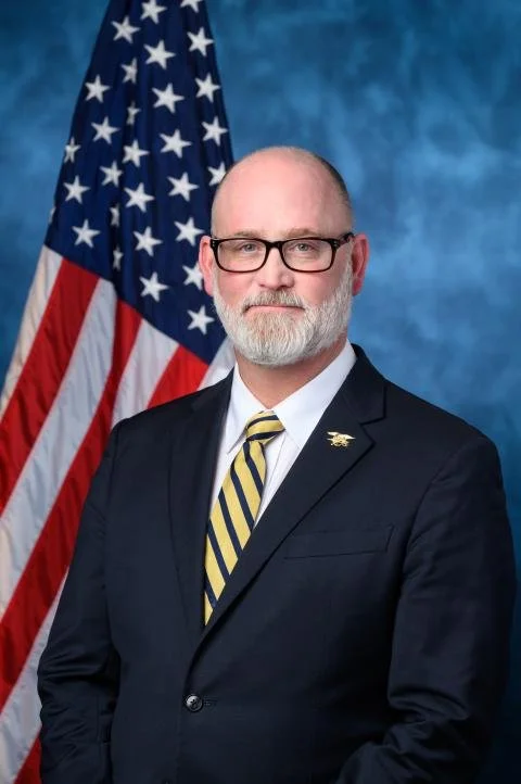 A middle-aged man with glasses, a beard, and a mustache, dressed in a dark suit with a striped yellow and black tie, standing in front of an American flag and a blue backdrop.