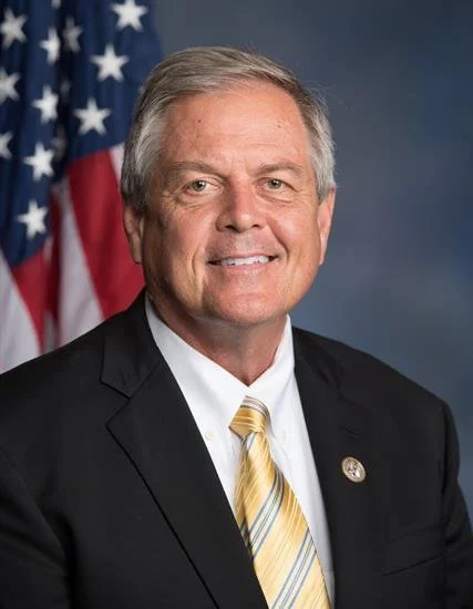 A smiling man in a suit posing in front of an American flag.