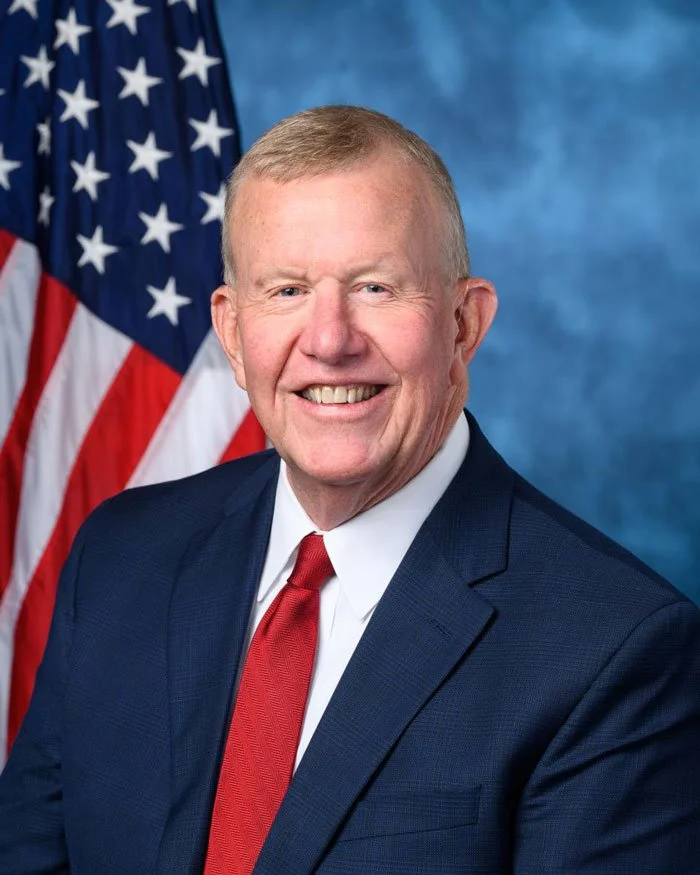 A smiling man in a dark suit, white shirt, and red tie, standing in front of an American flag with a blue background.