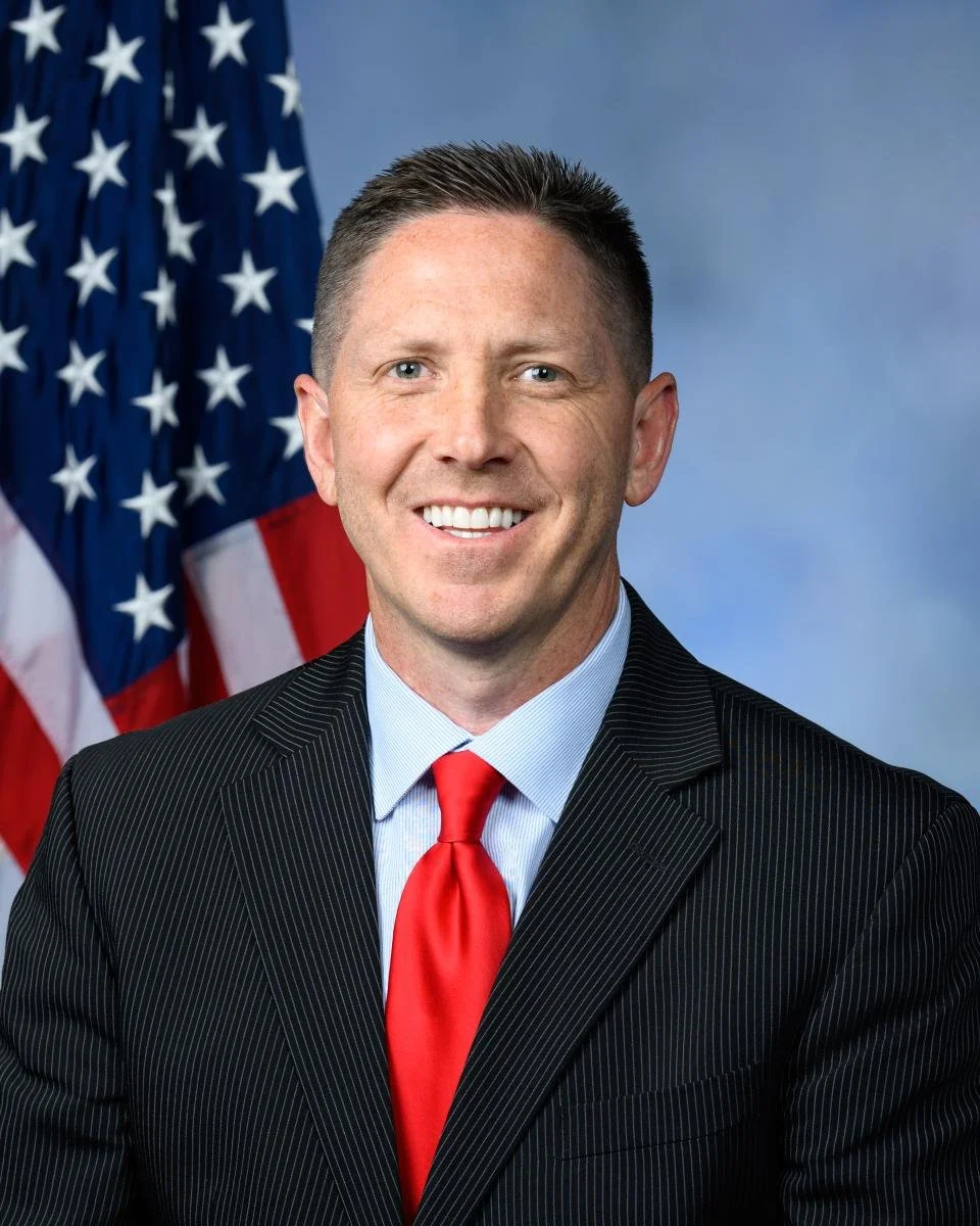 A smiling man in a suit and red tie, standing in front of an American flag.