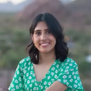 A woman with shoulder-length dark hair, smiling, wearing a green patterned top, standing outdoors with a blurred natural background.
