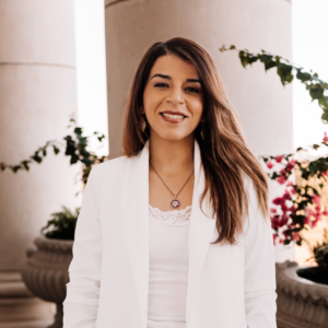 Young woman with long brown hair smiling, wearing a white blazer and standing outdoors with plants and flowers in the background.