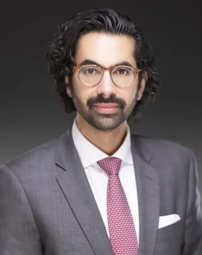 Portrait of a man with glasses, dark curly hair, wearing a gray suit, white shirt, pink patterned tie, and a white pocket square, against a dark background.