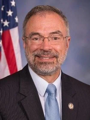 A man with glasses and a beard wearing a dark suit, white shirt, and light blue tie, standing in front of an American flag.