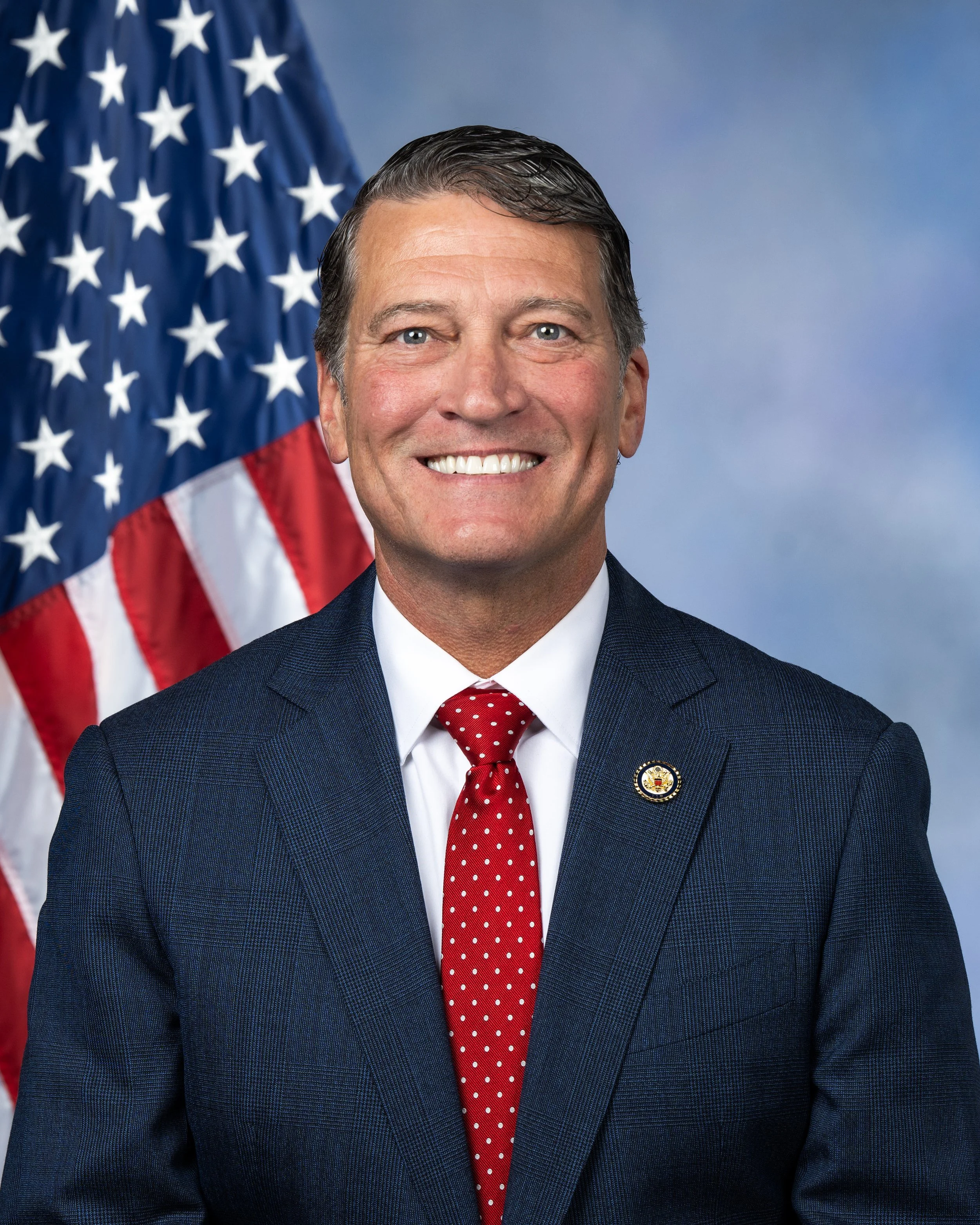 A smiling man in a dark blue suit, white shirt, and red polka dot tie, standing in front of an American flag.