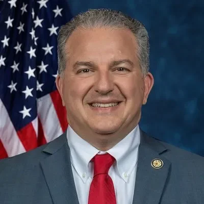 A man in a suit and red tie standing in front of the American flag, smiling at the camera.