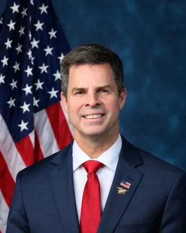 A man in a navy suit with a red tie and American flag pin, standing in front of an American flag backdrop.