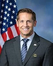 A man in a suit standing in front of an American flag, smiling at the camera.