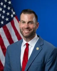 A smiling man with a beard, wearing a blue suit, white shirt, and red tie, standing in front of an American flag.