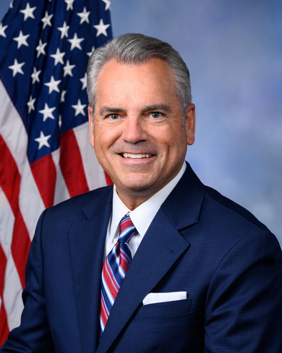 A man with gray hair and a bright smile, dressed in a navy blue suit, white shirt, and a red, white, and blue striped tie, posing in front of an American flag.