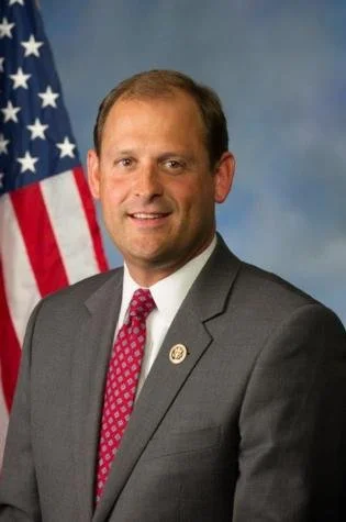 A man in a dark gray suit with a white shirt and red patterned tie stands in front of a U.S. flag, smiling at the camera.