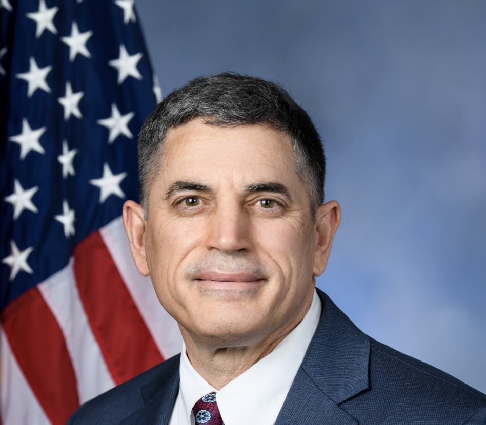 A middle-aged man in a suit and tie posing in front of the American flag with a blue background.