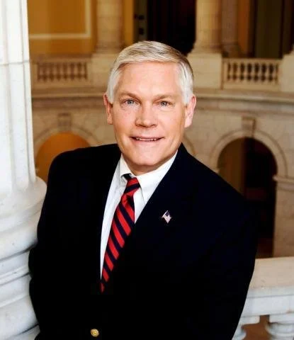 A smiling man in a suit and tie standing in front of an ornate building interior.