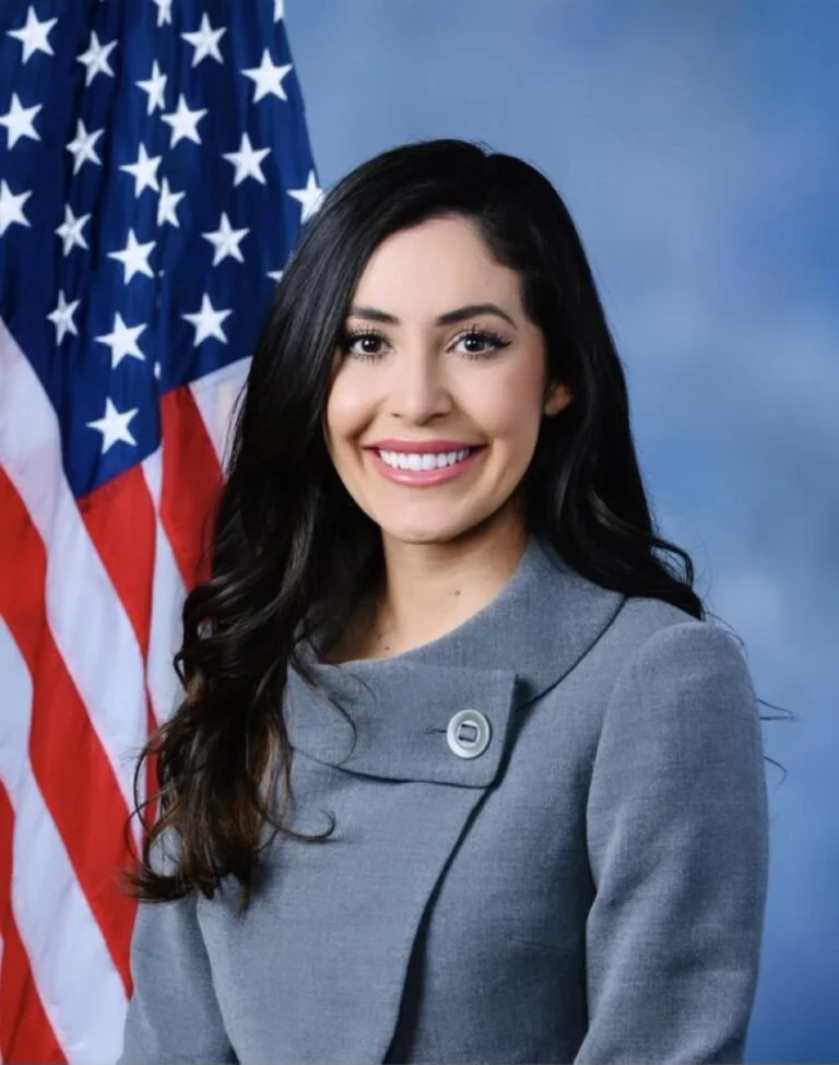 A woman with long dark hair smiling, dressed in a gray blazer, standing in front of a U.S. flag and a blue backdrop.