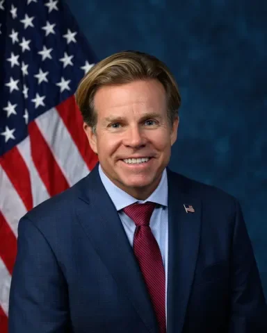 A man in a navy suit, light blue shirt, and red tie smiling for a portrait in front of an American flag.
