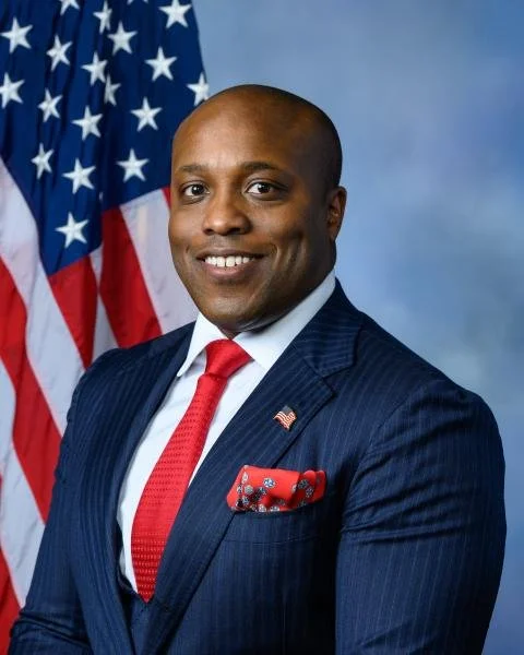 A man in a navy pinstripe suit with a red tie and pocket square, standing in front of an American flag with a blue background.
