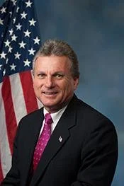 A man in a dark suit and purple tie standing in front of an American flag.