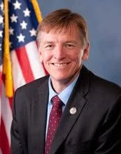 Portrait of a man in a suit, smiling, with an American flag in the background.