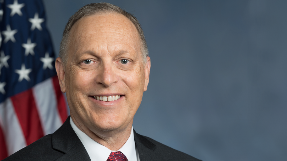 A man in a suit and red tie smiling in front of an American flag.