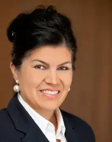 Portrait of a smiling woman with dark hair styled in an updo, wearing a navy blazer and pearl earrings.