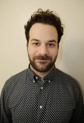 A man with dark, curly hair and a beard smiling at the camera, wearing a button-up, patterned shirt, standing against a plain, beige wall.