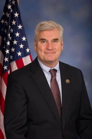 A man in a suit standing in front of an American flag with a dark blue background.