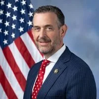 A man with dark hair and a beard, wearing a blue suit and red tie, standing in front of an American flag.