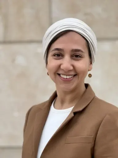 A woman smiling, wearing a white headscarf, tan blazer, and a white shirt, standing against a stone wall.