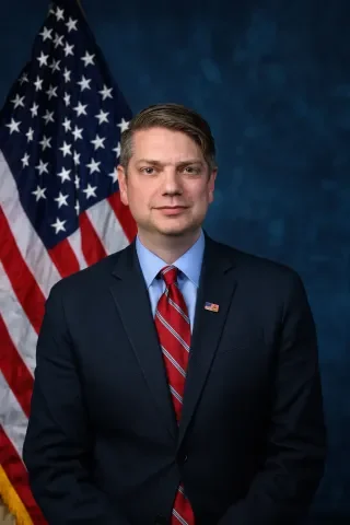 A man in a dark suit, light blue shirt, and red striped tie standing in front of an American flag with a dark blue background.