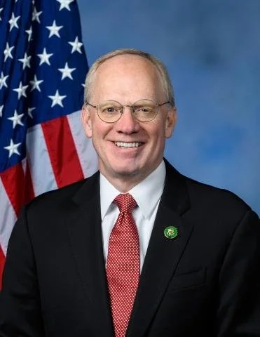 Portrait of a smiling man in a suit and tie, standing in front of an American flag.